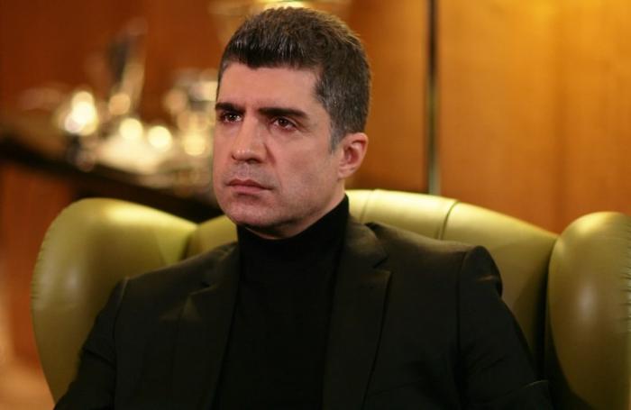 Özcan Deniz poartă o maletă neagră și un palton de aceeași culoare