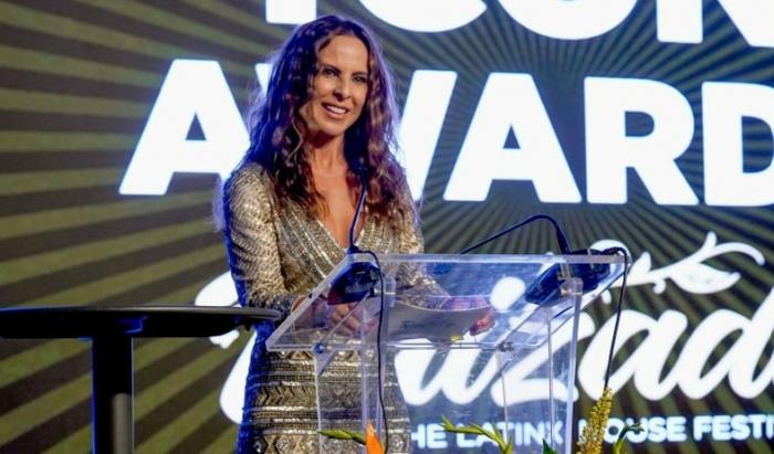 Kate del Castillo poartă o rochie cu mâneci lungi și stă pe scenă în fața unui microfon