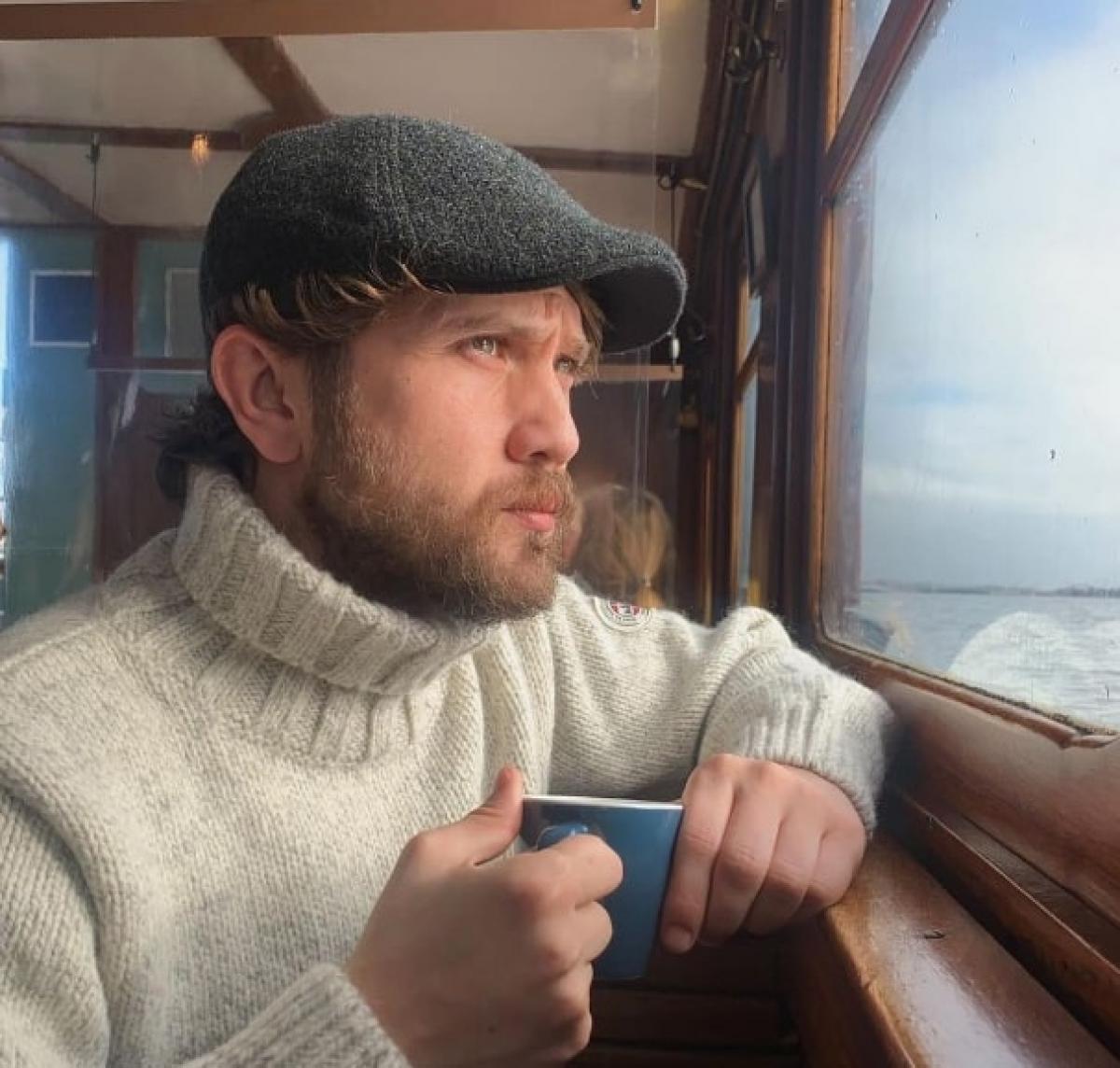 Aras Bulut Iynemli poartă un pulover deschis la culoare și ține o cană în mână