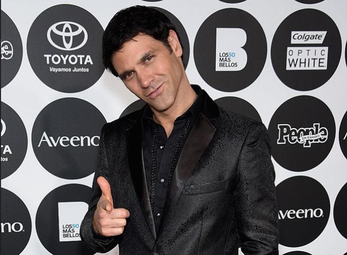 Valentino Lanús poartă o cămașă neagră și un sacou negru și zâmbește