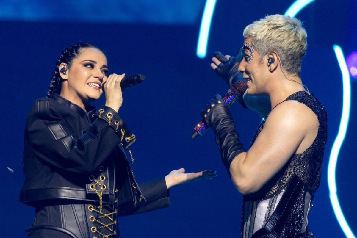 Maite Perroni poartă o ținută neagră și ține microfonul în mâna dreaptă, iar Christian Chávez poartă un maiou de aceeași culoare