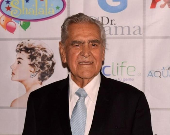 Eric del Castillo poartă un costum negru, o cămașă albă și o cravată și zâmbește