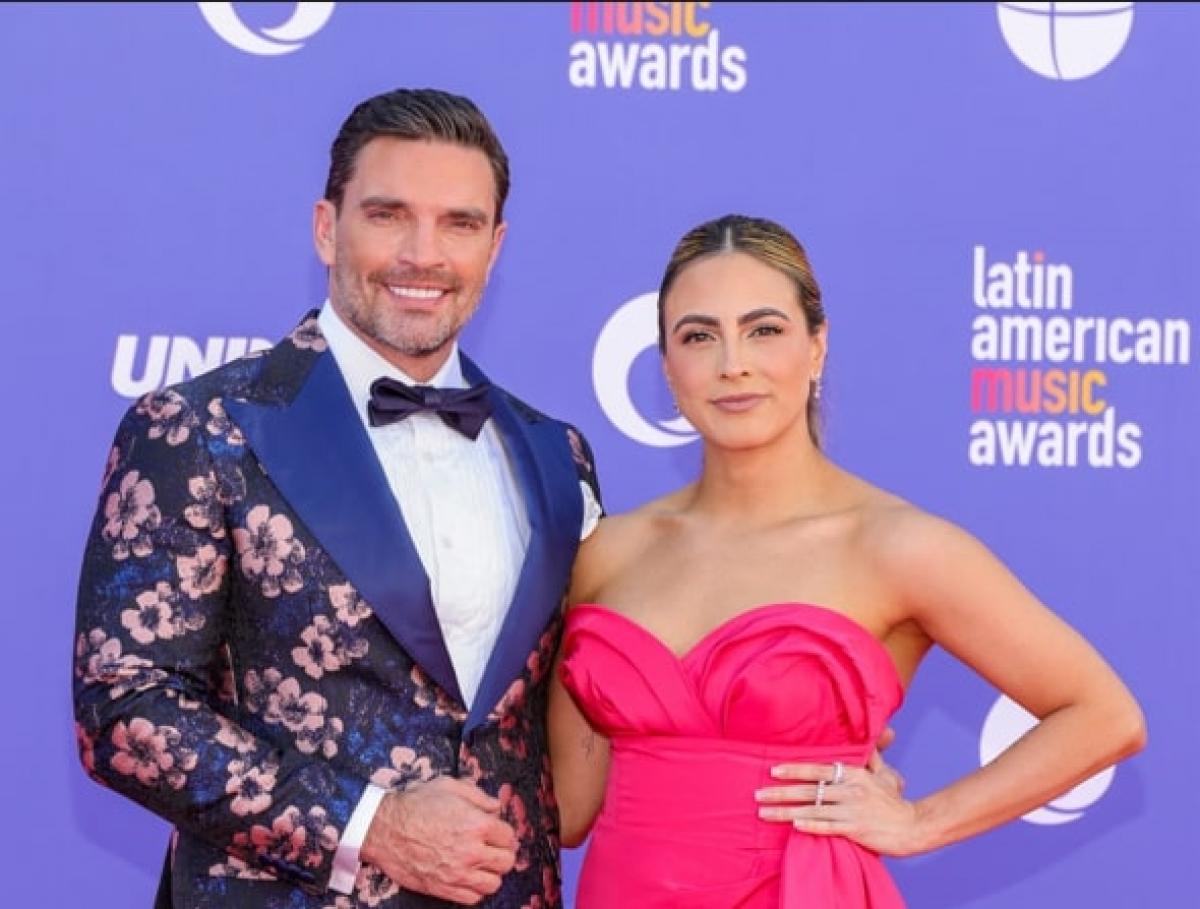 actor poartă un sacou cu imprimeu floral și stă lângă partenera sa care poartă o rochie roz fără mâneci