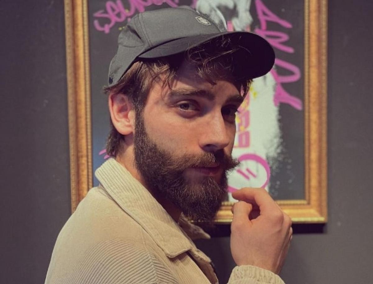 Burak Berkay Akgül poartă o șapcă și o jachetă maro deschis