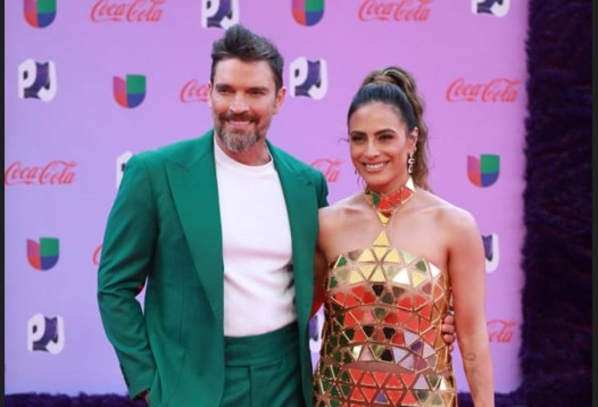 Julian Gil poartă un tricou alb și un costum verde și stă lângă Valeria Marin