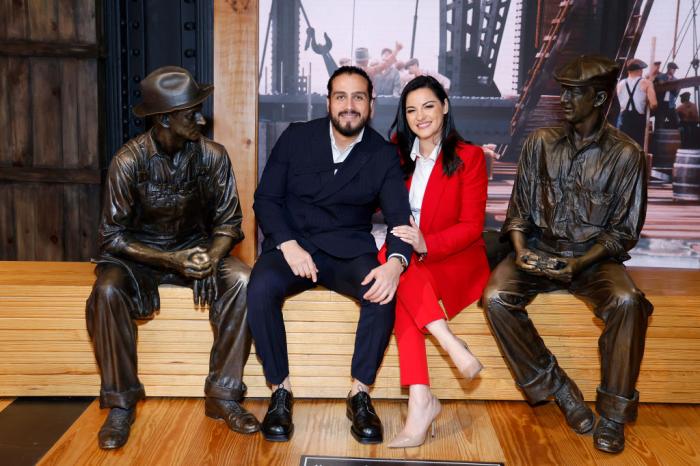 Maite Perroni poartă un costum roșu, pantofi cu toc și o cămașă albă și stă lângă partenerul său, Andrés Tovar