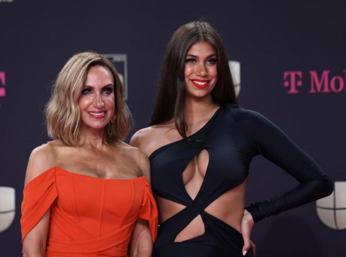 Lili Estefan poartă o rochie portocalie cu decolteu, zâmbește, are părul blond, desprins până la umeri și stă lângă fiica sa,Lina Teresa care poartă o rochie neagră, decupată
