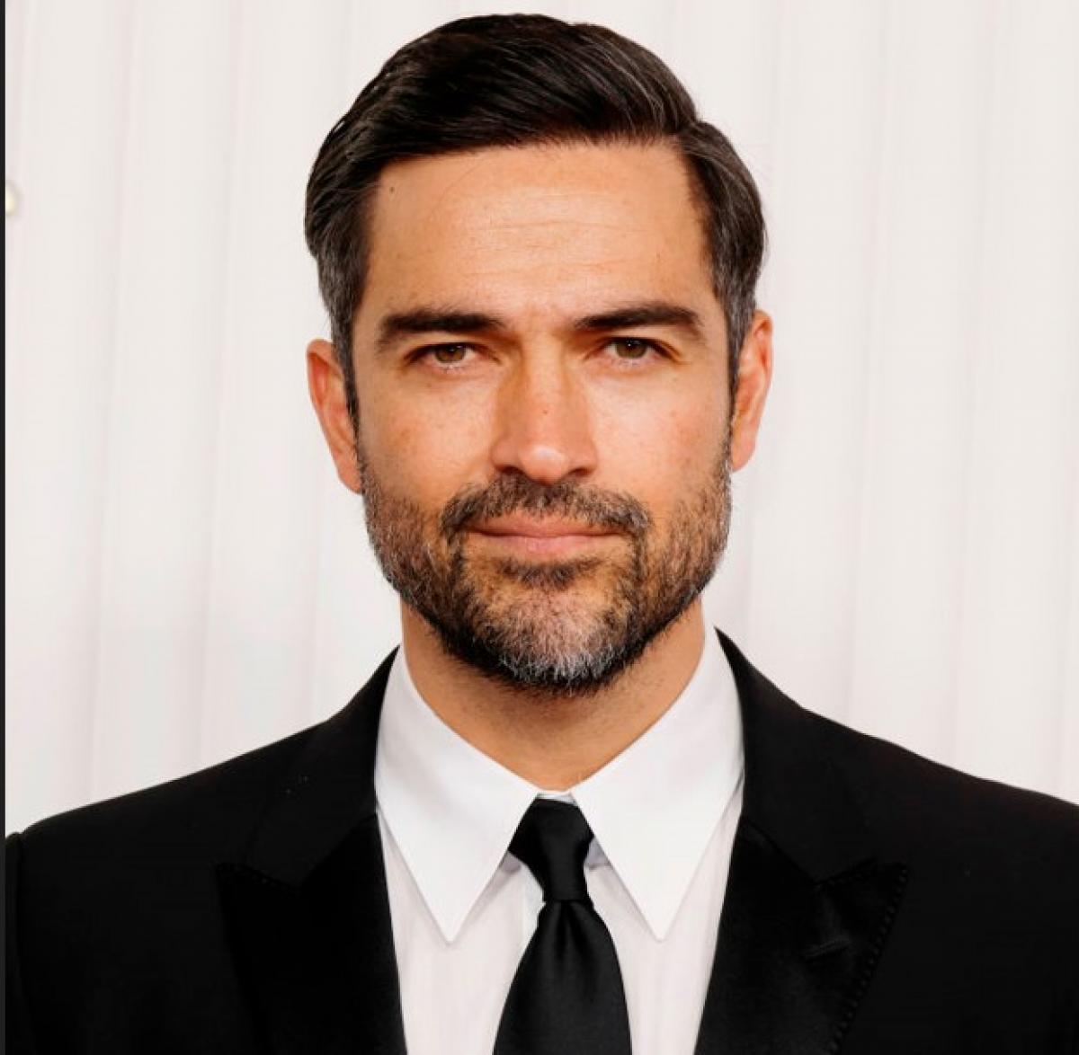 Alfonso Herrera poartă o cămașă albă, cravată neagră și sacou negru