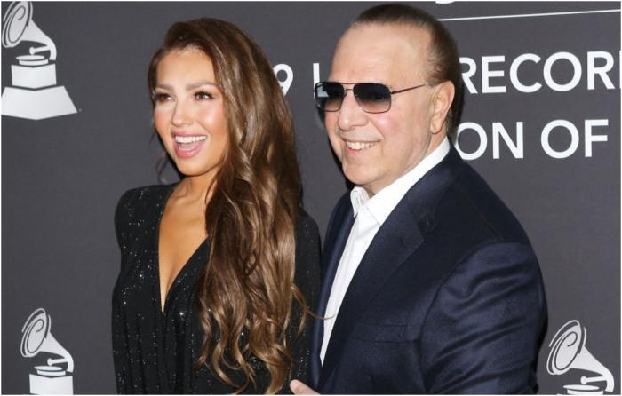 Thalía poartă o rochie neagră cu decolteu și zâmbește cu părul aranjat pe o parte și stă lângă Tommy Mottola