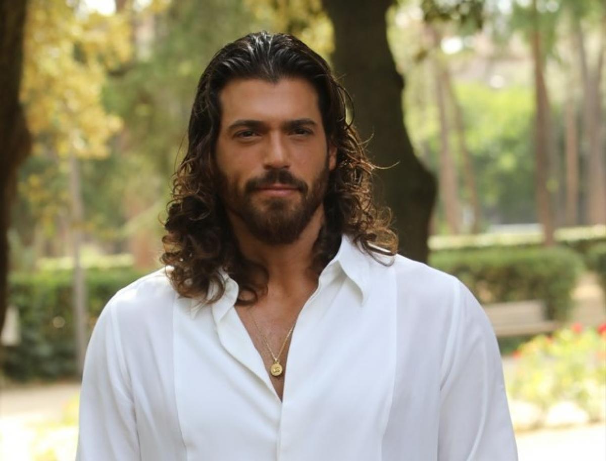 Can Yaman poartă o cămașă albă și are părul lung, ondulat