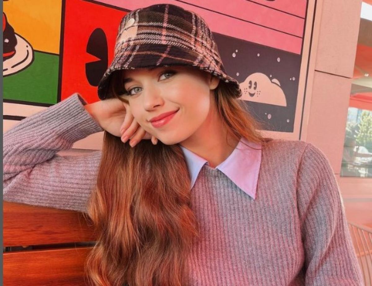 Afra Saraçoğlu poartă o cămașă albă și un pulover gri, o pălărie în carouri și părul lung, desprins