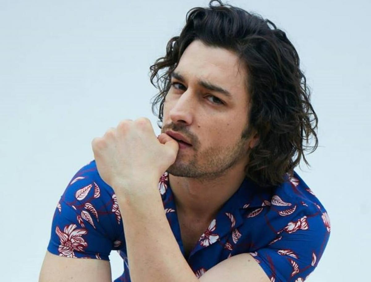 Alp Navruz poartă o cămașă albastră cu flori și își ține mână stângă aproape de gură