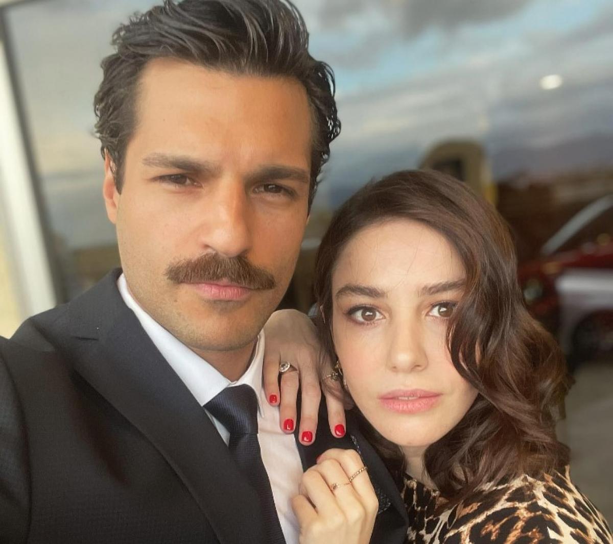 Serkan Çayoğlu poartă o cămașă albă și un sacou negru și stă lângă Özge Gürel