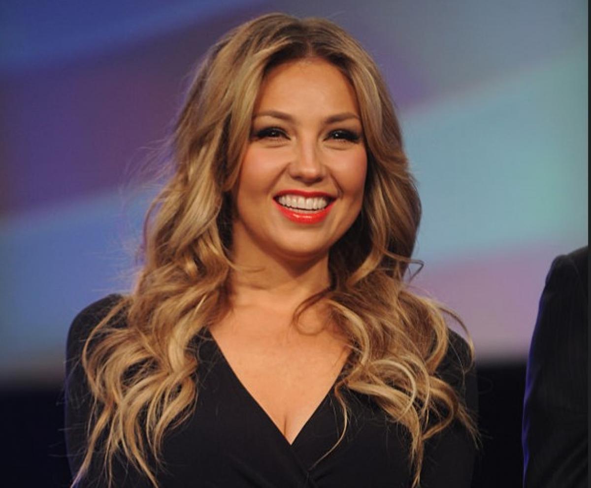 Thalía poartă o rochie neagră cu decolteu și zâmbește, are părul lung, desprins