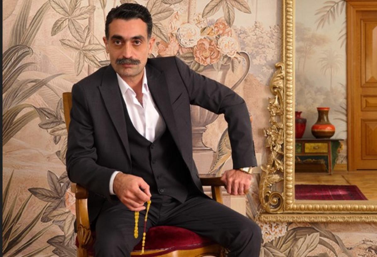 Diren Polatoğulları poartă o cămașă albă și un costum negru și stă pe scaun ținând în mână o cruce