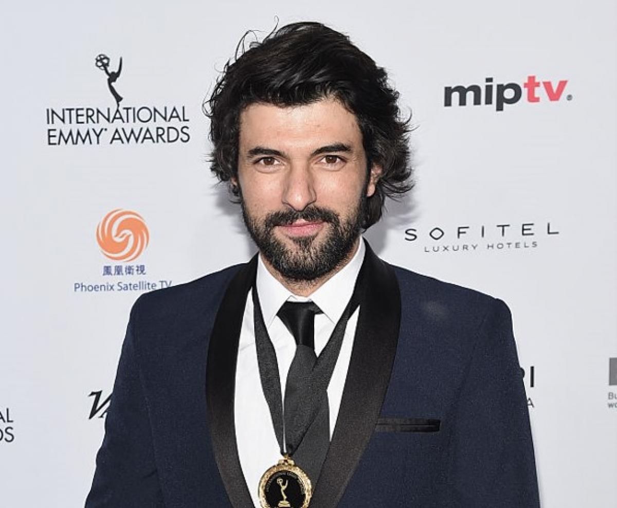 Engin Akyürek poartă un costum negru și o cămașă albă și are părul lung