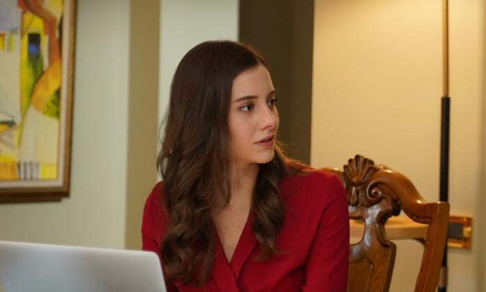 Beril Pozam poartă o rochie vișinie cu mâneci lungi și stă pe scaun cu un laptop în față
