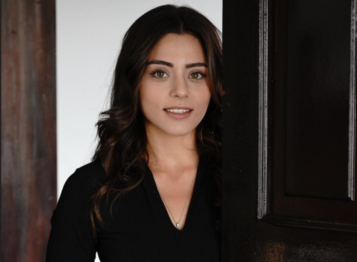 Sıla Türkoğlu poartă o bluză neagră cu mâneci lungi și decolteu și zâmbește