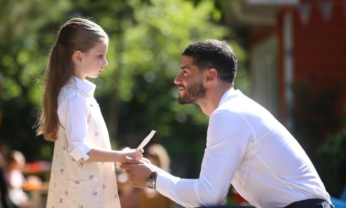 Berk Oktay poartă o cămașă albă și stă în fața micuței Azra Aksu care poartă o rochie cu flori