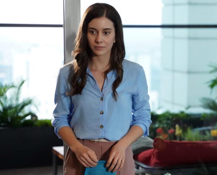 Beril Pozam poartă o cămașă albastră cu mâneci lungi, are părul lung, desprins, ondulat și ține în mână o mapă