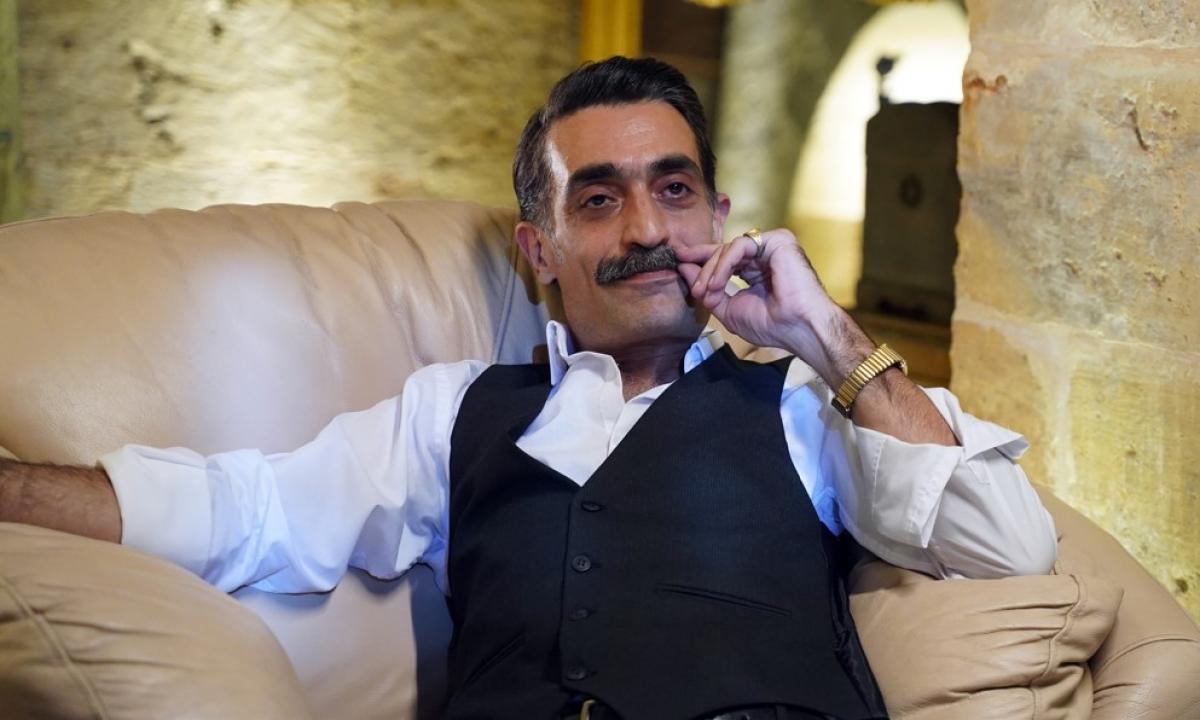 Diren Polatoğulları poartă o cămașă deschisă la culoare, un sacou negru