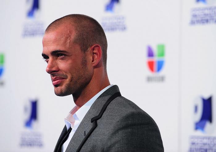 William Levy poartă o cămașă albă, un sacou gri și este chel
