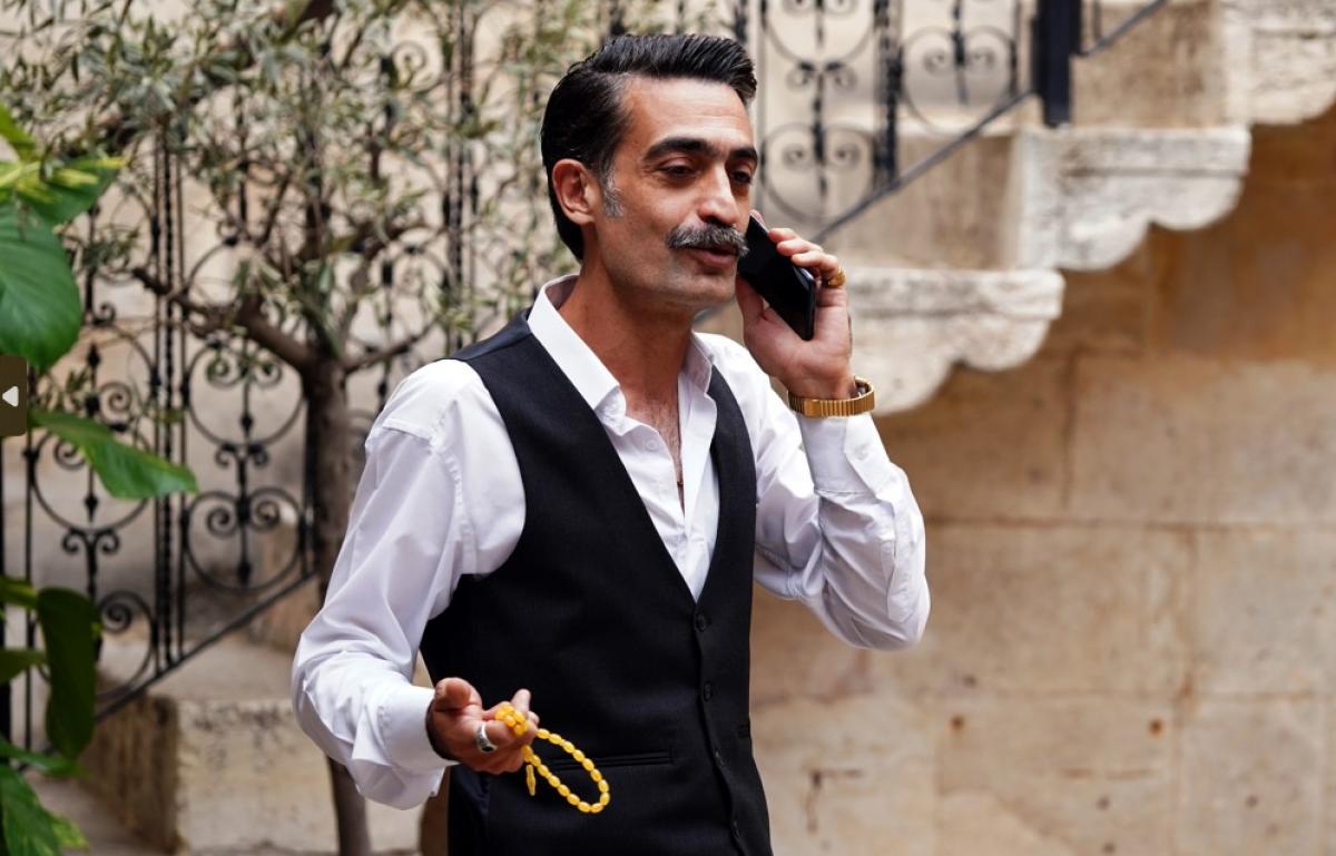 Diren Polatoğulları poartă o cămașă albă și o vestă neagră și vorbește la telefon