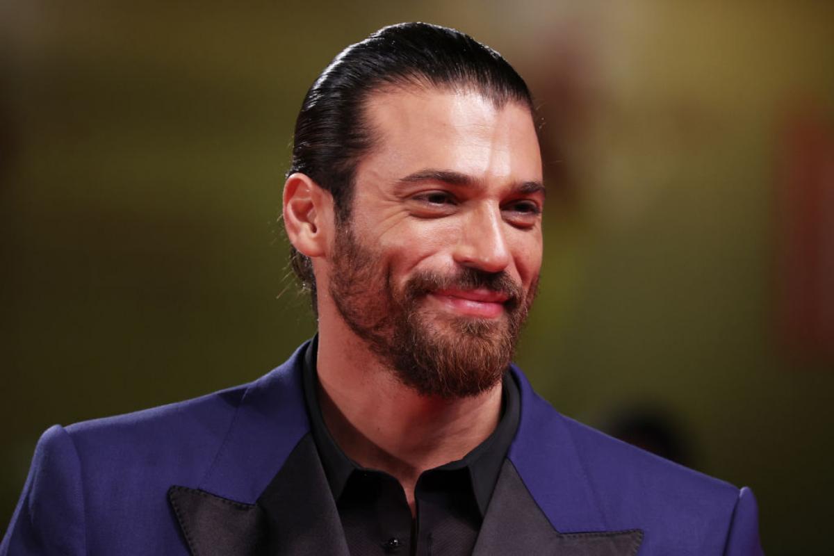 Can Yaman poartă o cămașă neagră și un palton albastru și zâmbește