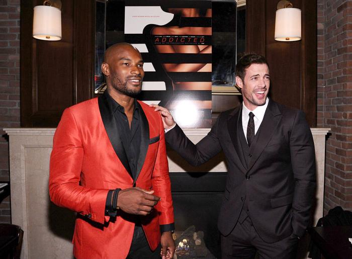 Tyson Beckford poartă un costum portocaliu și zâmbește, iar William Levy poartă un costum negru și își ține mâna dreaptă pe umărul colegului