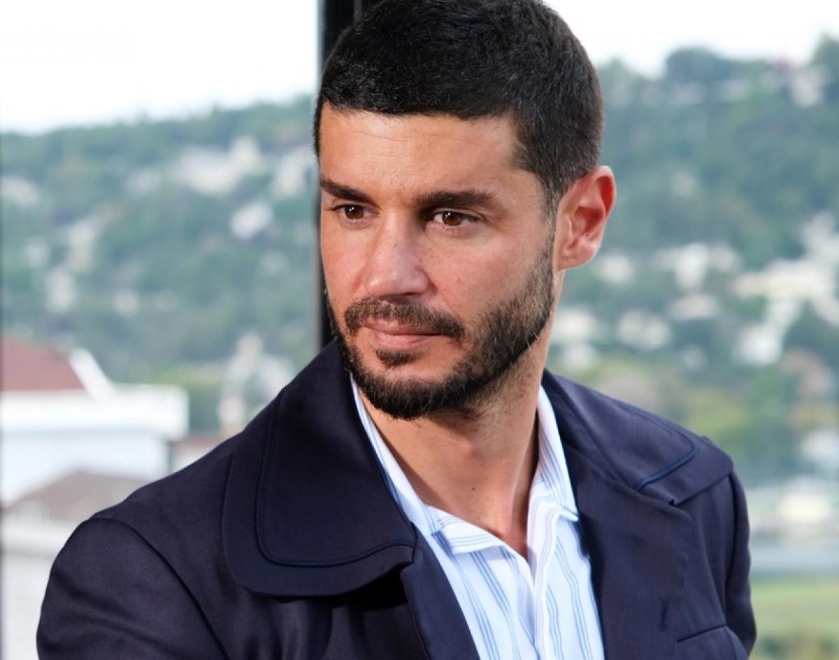 Berk Oktay poartă o cămașă albastră și un sacou închis la culoare