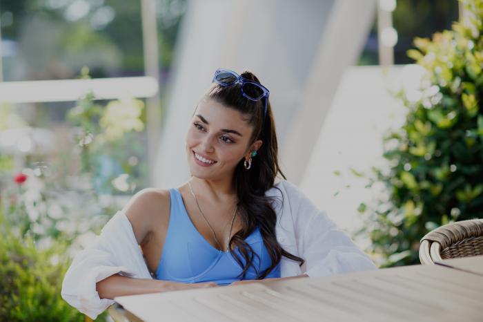 Hande Erçel poartă un maiou albastru și o bluză albă și are părul coafat ondulat