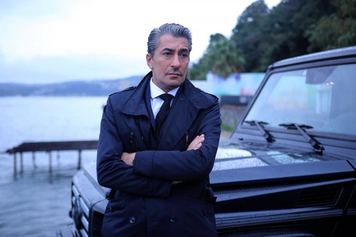 Erkan Petekkaya poartă o ținută all-black și stă sprijinit cu spatele de o mașină