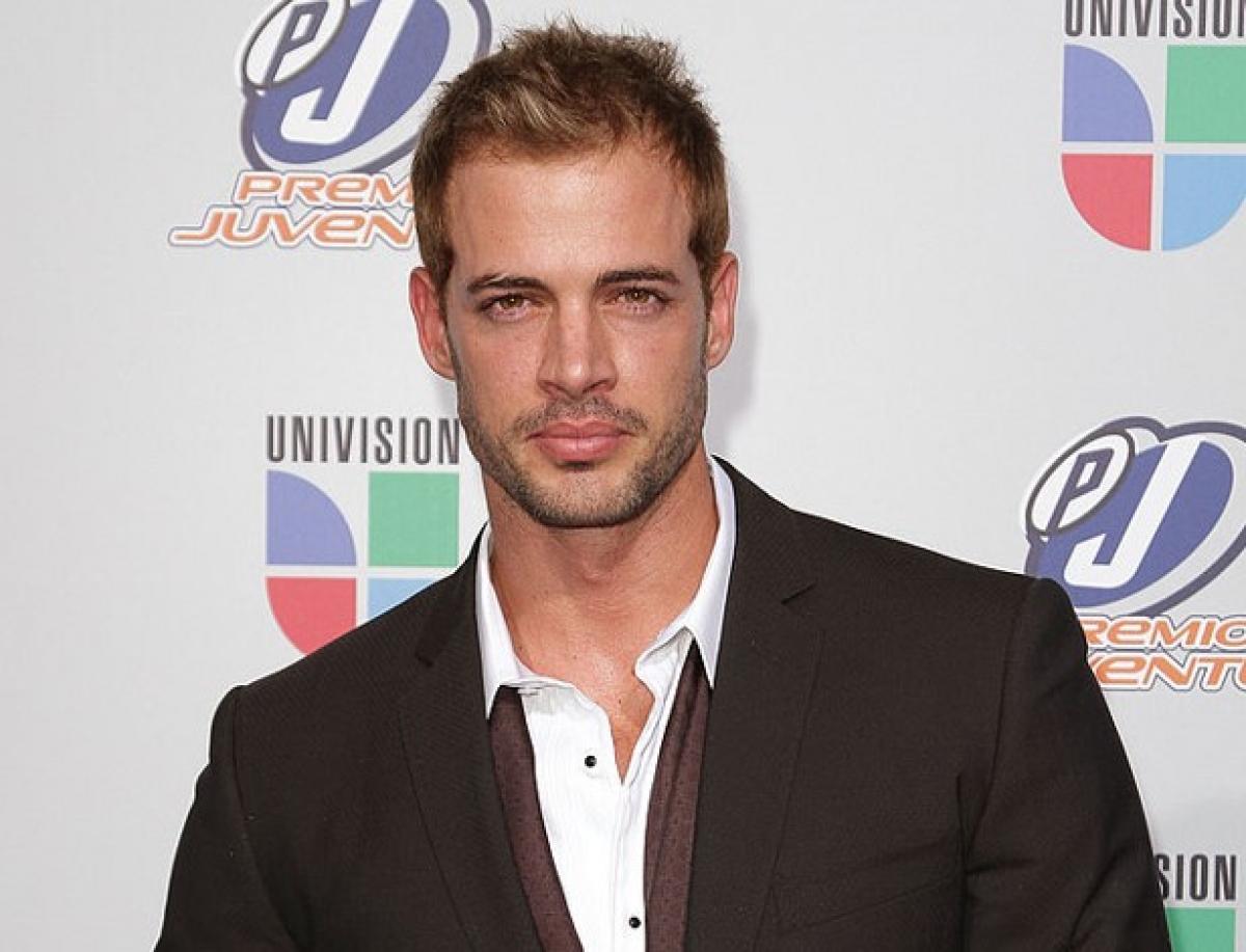 William Levy poartă o cămașă albă și un sacou negru cu mâneci lungi și zâmbește