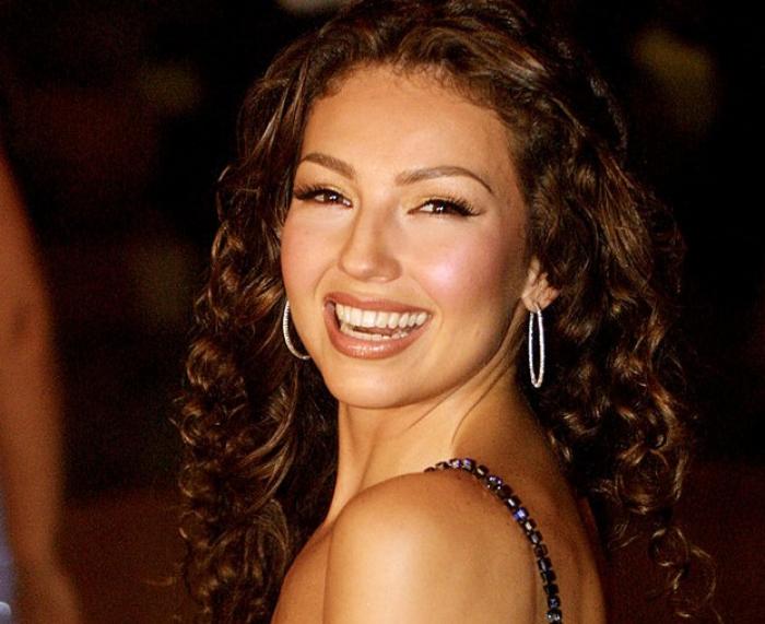 Thalía poartă părul lung și ondulat și zâmbește