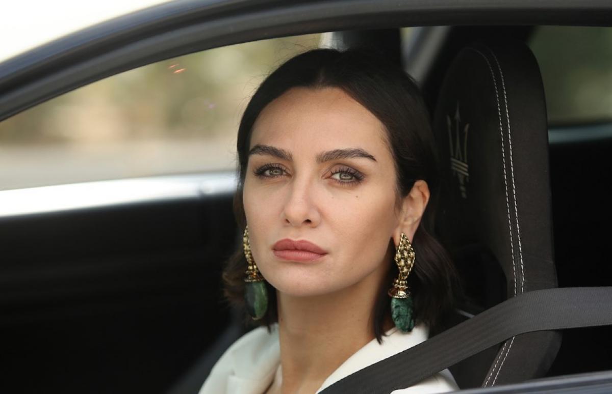 Birce Akalay poartă o bluză albă cu părul lung desprins, se află într-o mașină la volan