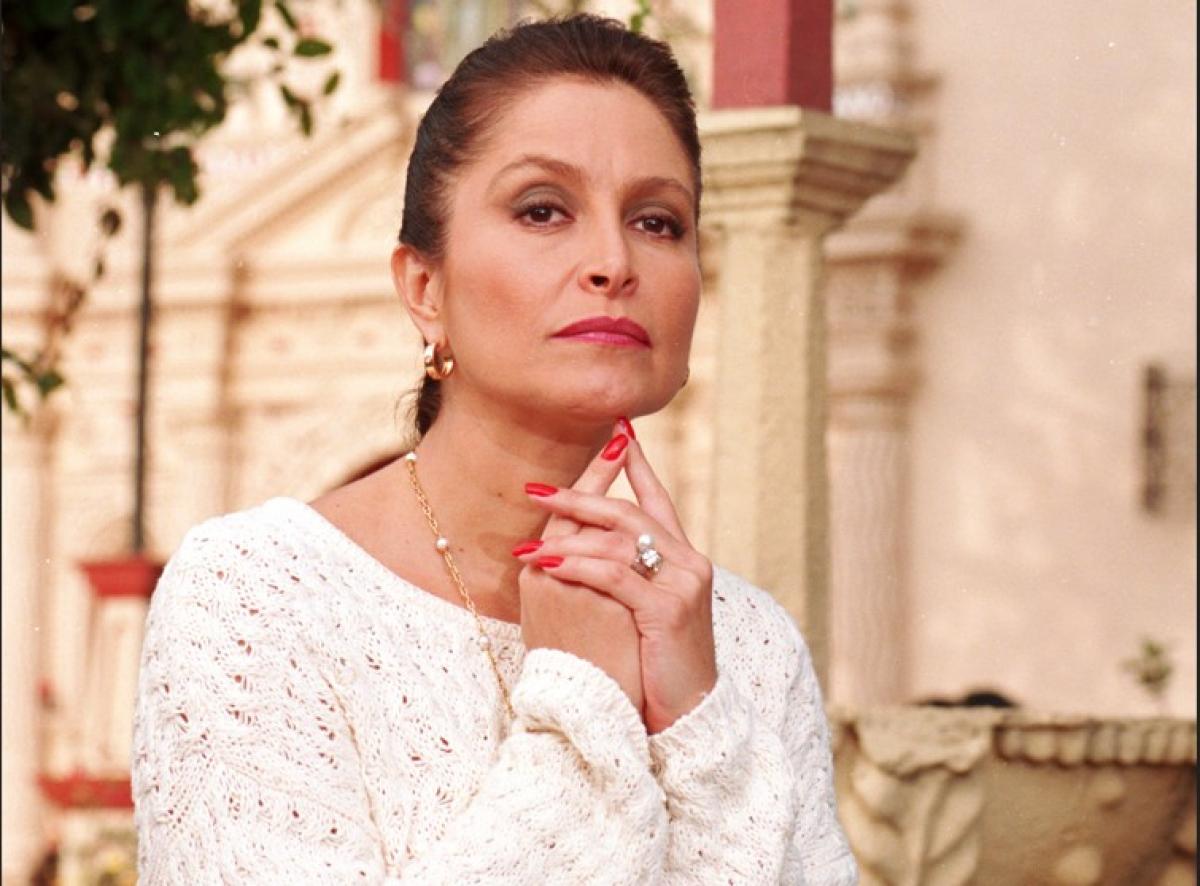 Daniela Romo poartă o bluză albă cu mâneci lungi și își ține mâinile împreunate