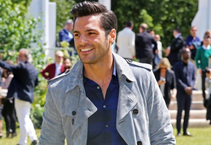 Serkan Çayoğlu poartă un tricou albastru și o jachetă lungă și zâmbește