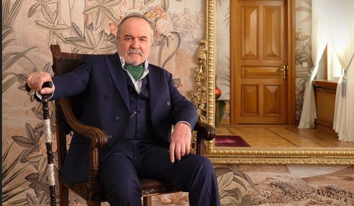 Çetin Tekindor poartă un costum închis la culoare și stă pe scaun