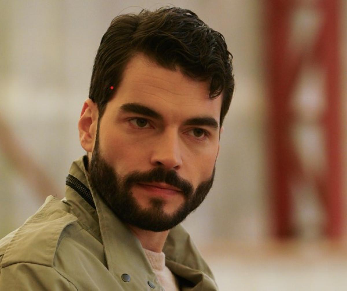 Akin Akinozu poartă o geacă deschisă la culoare și are barbă