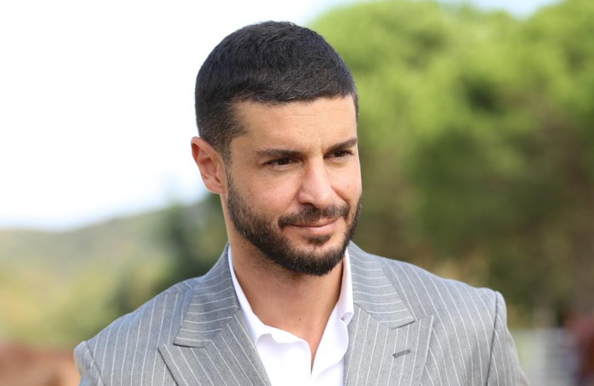 Berk Oktay poartă o cămașă albă și un sacou gri