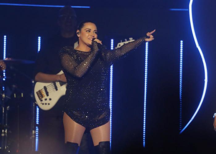 Maite Perroni poartă o ținută neagră și ține microfonul în mâna dreaptă
