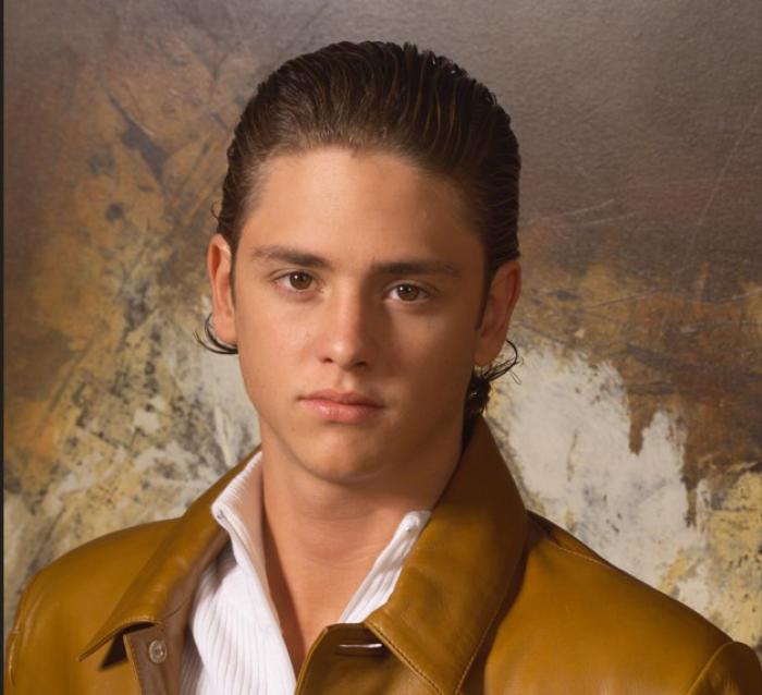 Christopher Uckermann poartă o cămașă albă și o geacă maro de piele