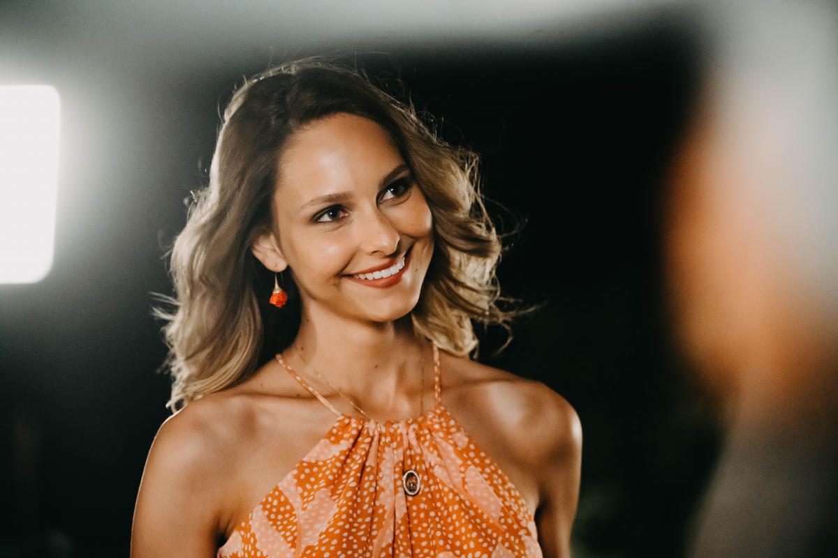 Jessica May poartă o bluză portocalie fără mâneci și zâmbește