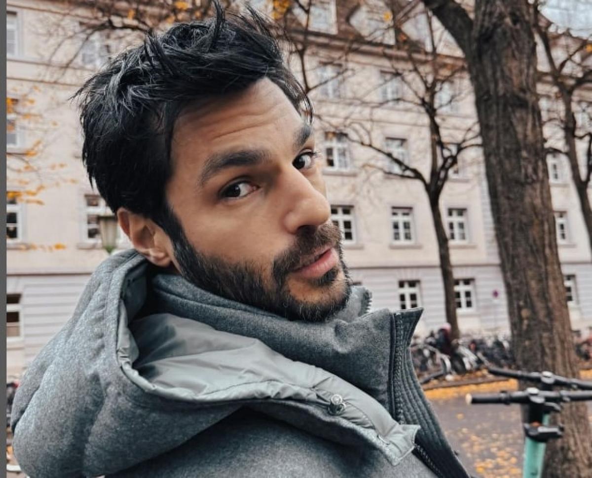 actor poartă o geacă cu glugă și privește spre cameră