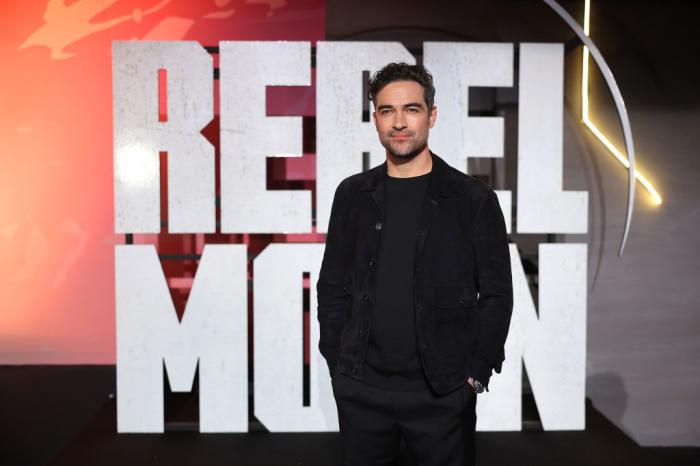 actor poartă un costum negru și în spatele său stă scris numele Rebel Moon