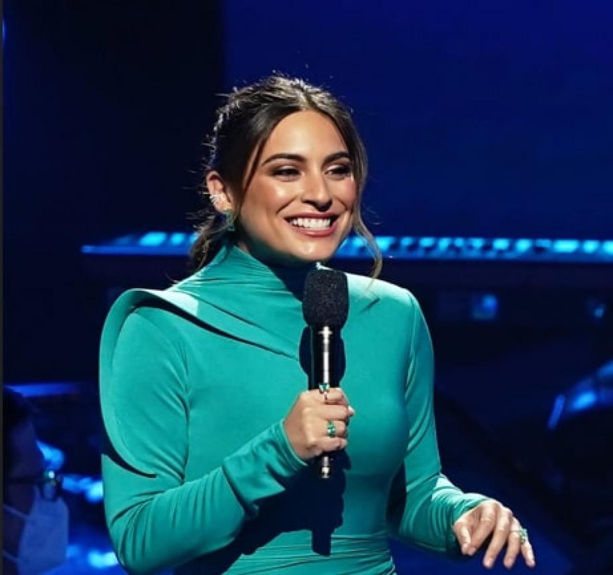 Ana Brenda Contreras ține un microfon în mână și zâmbește pe un fundal închis la culoare