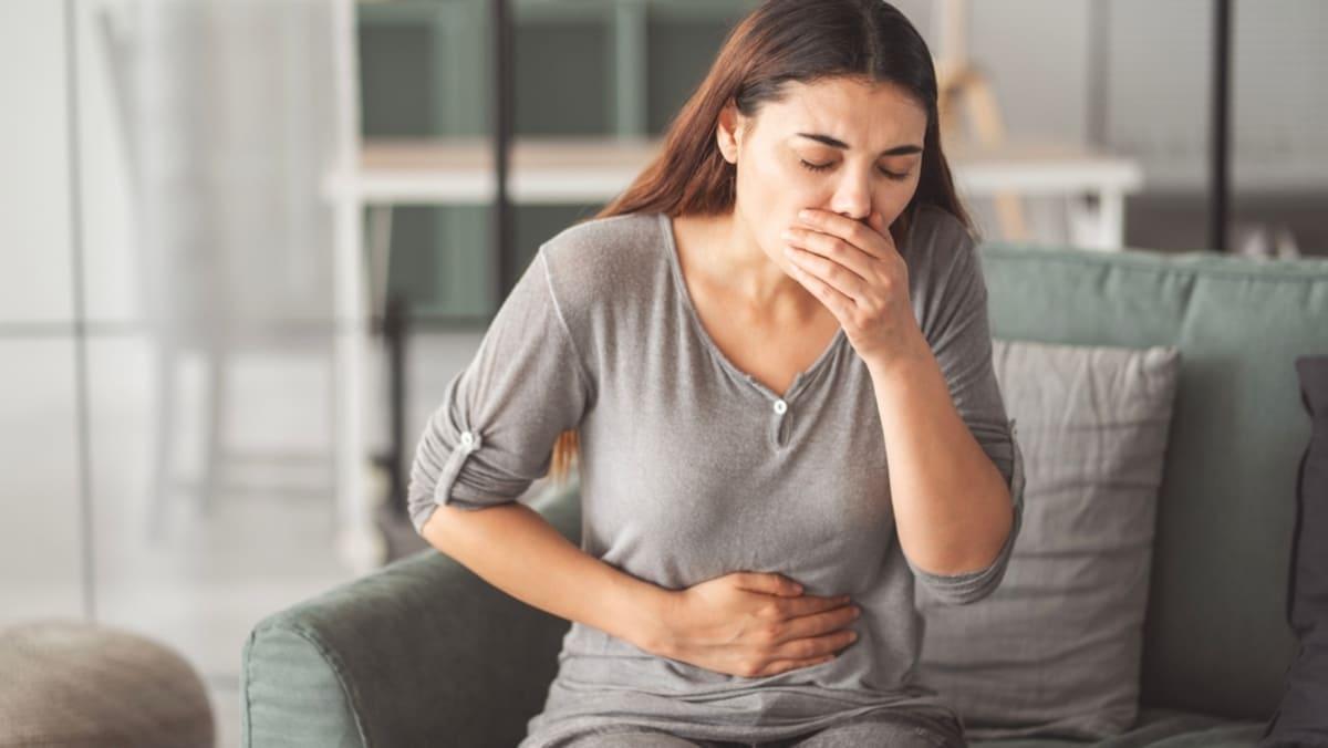 O femeie cu tricou gri și părul lung, ține o mână la gură și una pe abdomen