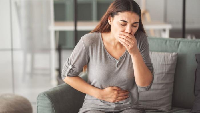 (P) Greața și vărsăturile: cauze frecvente și momentele în care vizita la medic devine necesară O femeie cu tricou gri și părul lung, ține o mână la gură și una pe abdomen