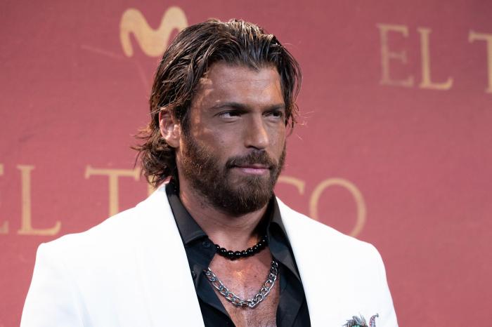 Can Yaman poartă un sacou alb și o cămașă neagră pe un fundal deschis la culoare