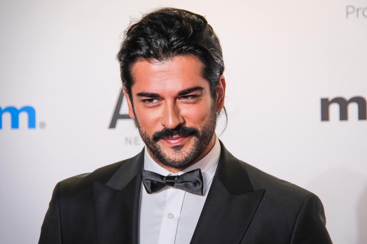 Burak Ozcivit poartă o cămașă albă și sacou negru pe un fundal deschis la culoare